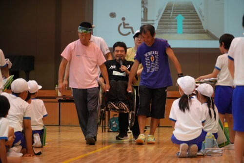 先生4人が講師を車いすごと持ち上げている場面